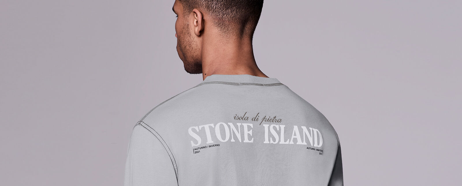 Close up shot of model showing the back of a light gray shirt with contrast dark stitching and lettering that reads isola di pietra STONE ISLAND AUTUNNO / INVERNO 2021 AUTUMN / WINTER 2021.