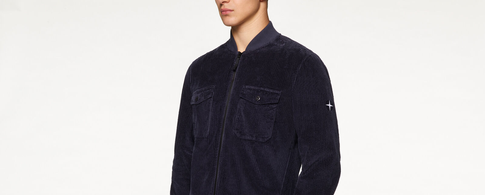 Close up shot of model wearing a dark blue corduroy bomber jacket with zipper fastening, two chest pockets with flap and button and the two-tone blue and white Stone Island Compass Rose embroidery on the upper left sleeve.