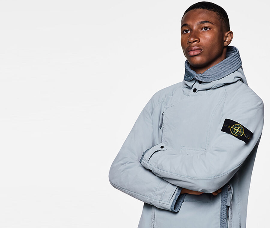 Close up shot of model wearing a light gray jacket with standing collar, diagonal two way zipper fastening, strap at cuffs held by a snap and the Stone Island badge on the upper left sleeve.