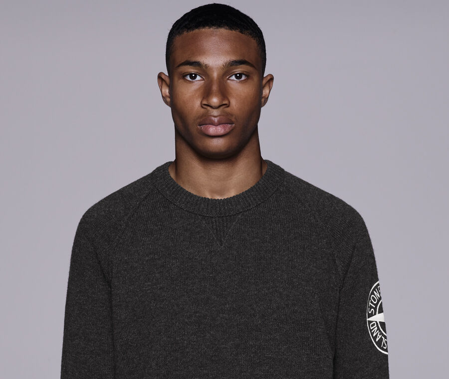 Model wearing a dark colored mélange crewneck sweater with raglan sleeves, white embroidery of the Stone Island Compass logo on the left sleeve and ribbed neckline, cuffs, and bottom band.