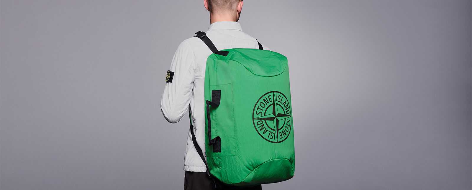 Model, shot from behind, wearing an off-white shirt with the Stone Island badge on the upper left sleeve and a large green travel backpack with the Stone Island compass rose logo.