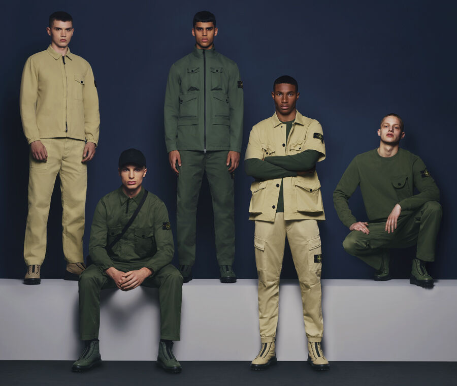 Five models set against a navy blue backdrop. Three wearing various dark green jackets, sweaters, pants and boots; one wearing a khaki jacket, pants and boots and the other wearing a dark green sweatshirt combined with a short-sleeved khaki jacket, pants