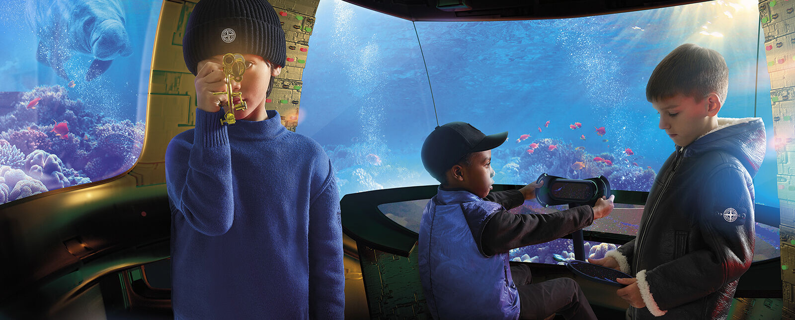 In the water, three junior models inside a deep sea shuttle, one wearing a blue sweater and black ribbed beanie, the second one a blue vest and a black cap and the third one a sheepskin jacket with hood and white lining.