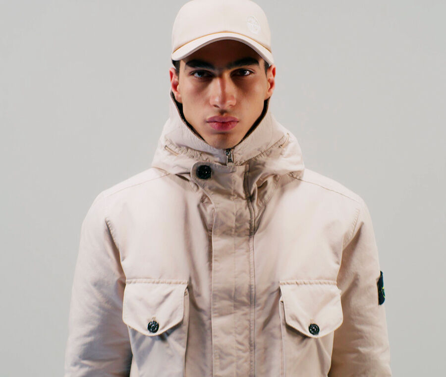Close up shot of model wearing an off white cap and a matching jacket with hood, central fastening hidden by a flap and two chest pockets with flap and button fastening.