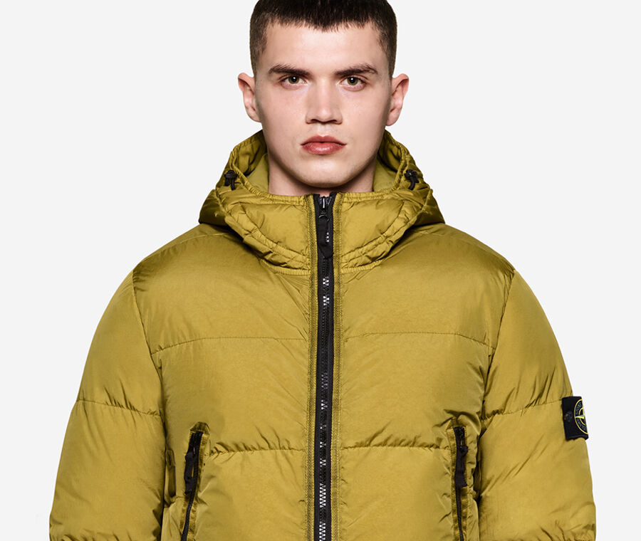 Model wearing dark colored pants and a mustard down jacket with hood, black zipper fastening, two side pockets with zipper and the Stone Island badge on the upper left sleeve.
