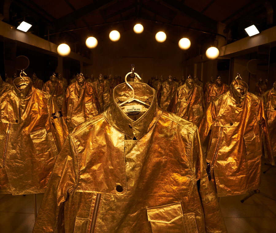 Numerous rows of Stone Island Prototype Research Series 05 Copper Nanotechnology jackets displayed in a warehouse setting under bright lighting.