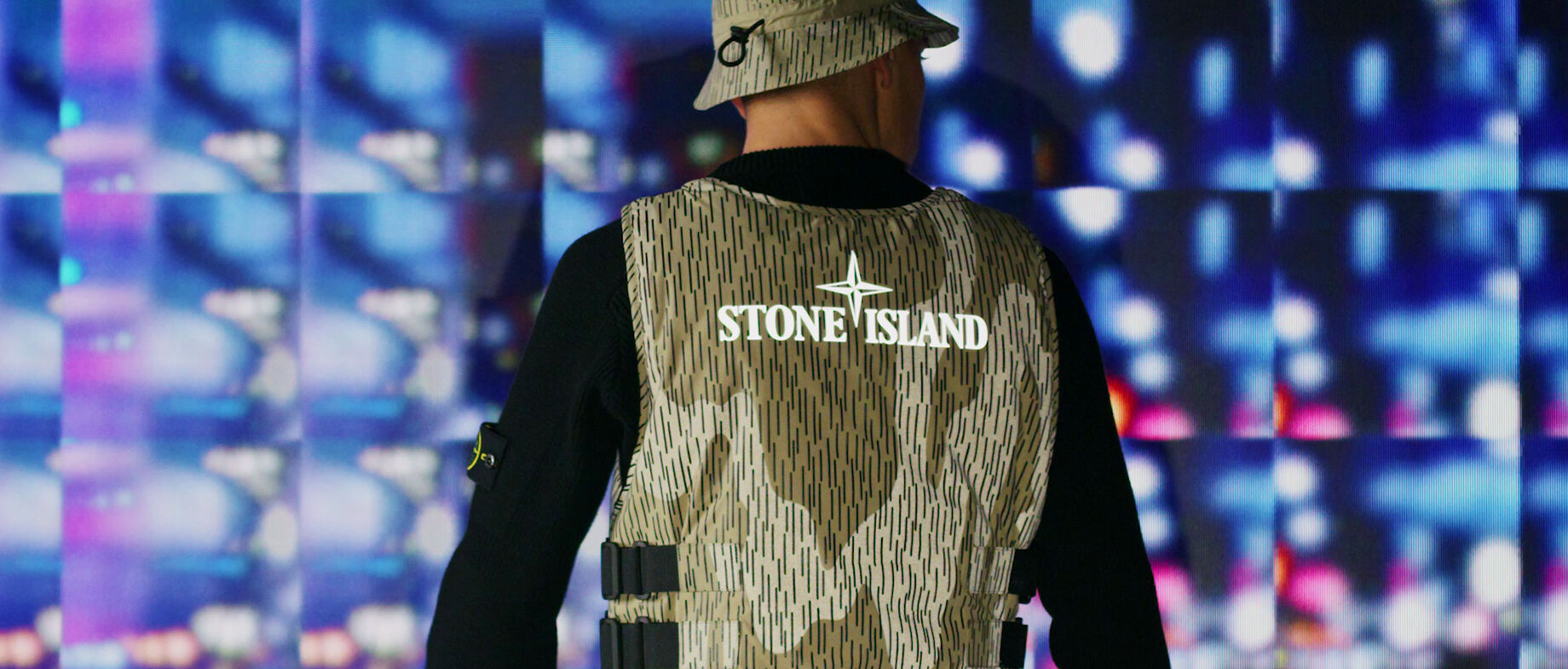 Model, shot from behind, wearing a black sweater; khaki vertical line and reflective camo print bucket hat and vest featuring a Stone Island graphic logo.
