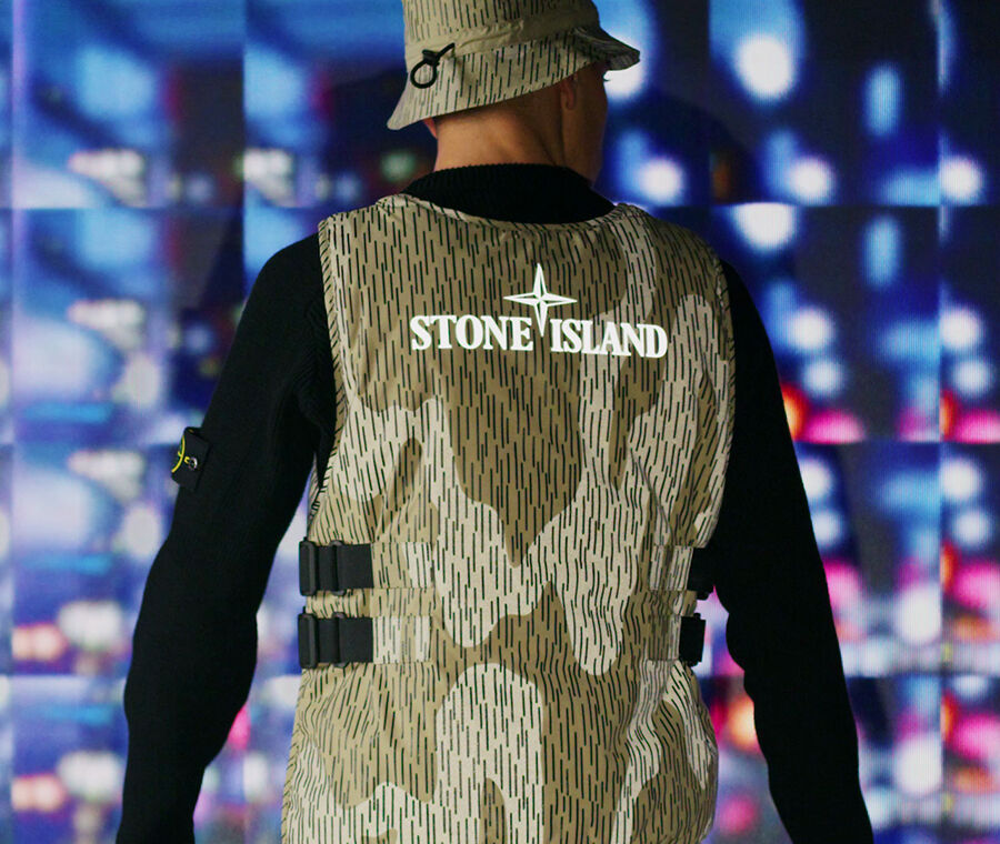 Model, shot from behind, wearing a black sweater; khaki vertical line and reflective camo print bucket hat and vest featuring a Stone Island graphic logo.