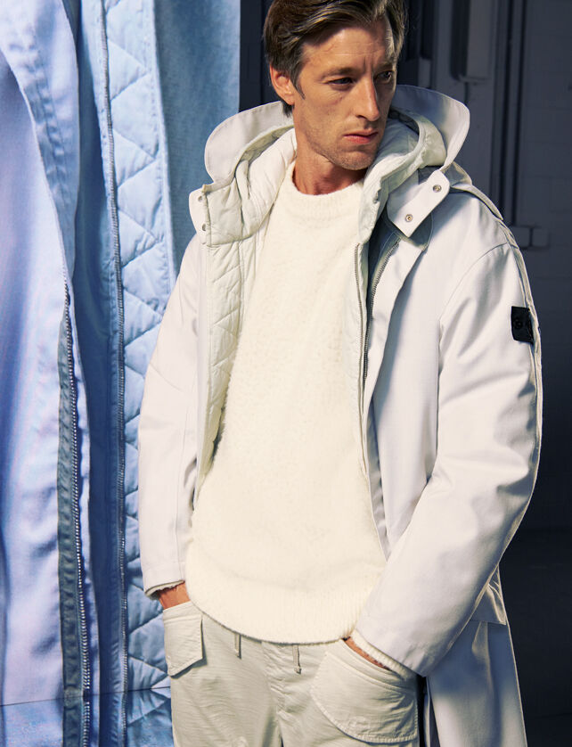 Model wearing a white long jacket, off-white sweater and pants, beside a tall screen with a close-up of the outfit.