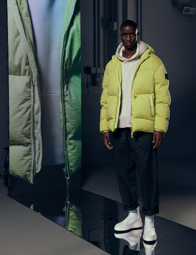 Model wearing a bright green padded jacket, off-white hoodie, black pants and white boots, beside a tall screen with a close-up of the outfit.