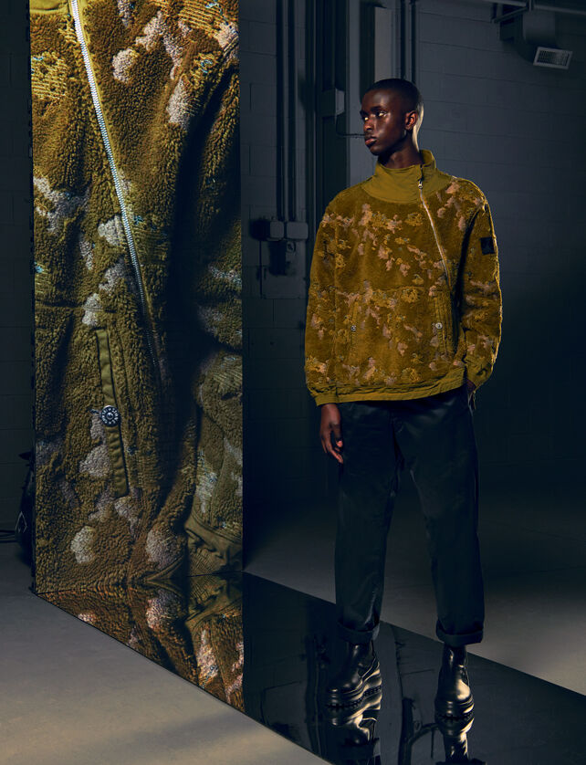 Model wearing a yellow fleece jacket, black pants and black boots, beside a tall screen with a close-up of the outfit.