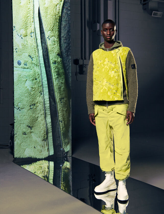 Model wearing a fluorescent yellow fleece jacket, a brown hoodie, yellow pants and white boots, beside a tall screen with a close-up of the outfit.