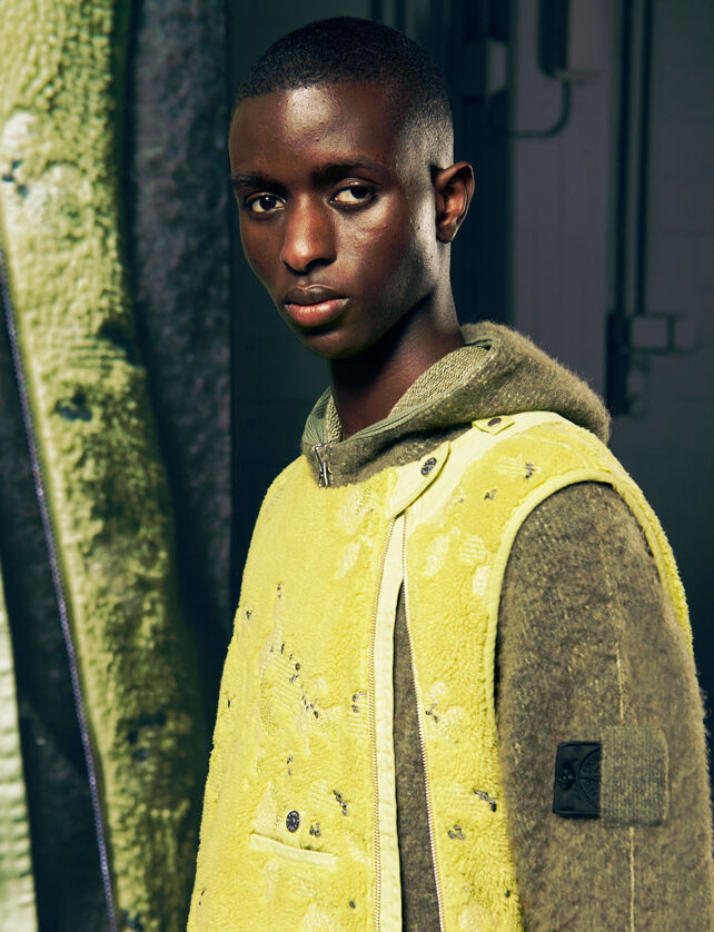 Model wearing a fluorescent yellow fleece jacket and a brown hoodie, beside a tall screen with a close-up of the outfit.
