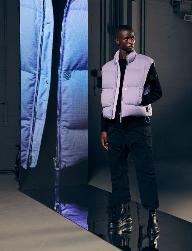 Model wearing a lilac padded vest jacket, a black sweater, pants and boots, beside a tall screen with a close-up of the outfit.