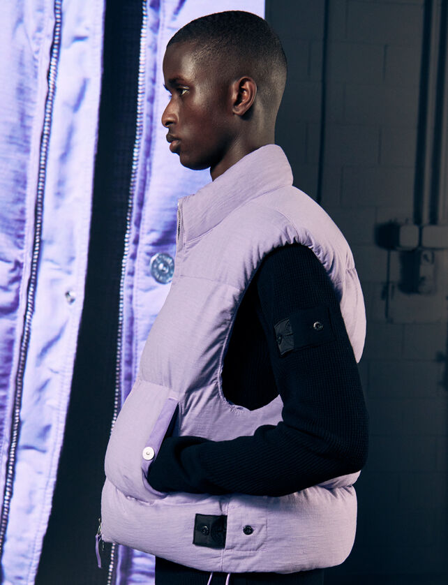 Model wearing a lilac padded vest jacket, and a black sweater with the Shadow Project badge on the sleeve.