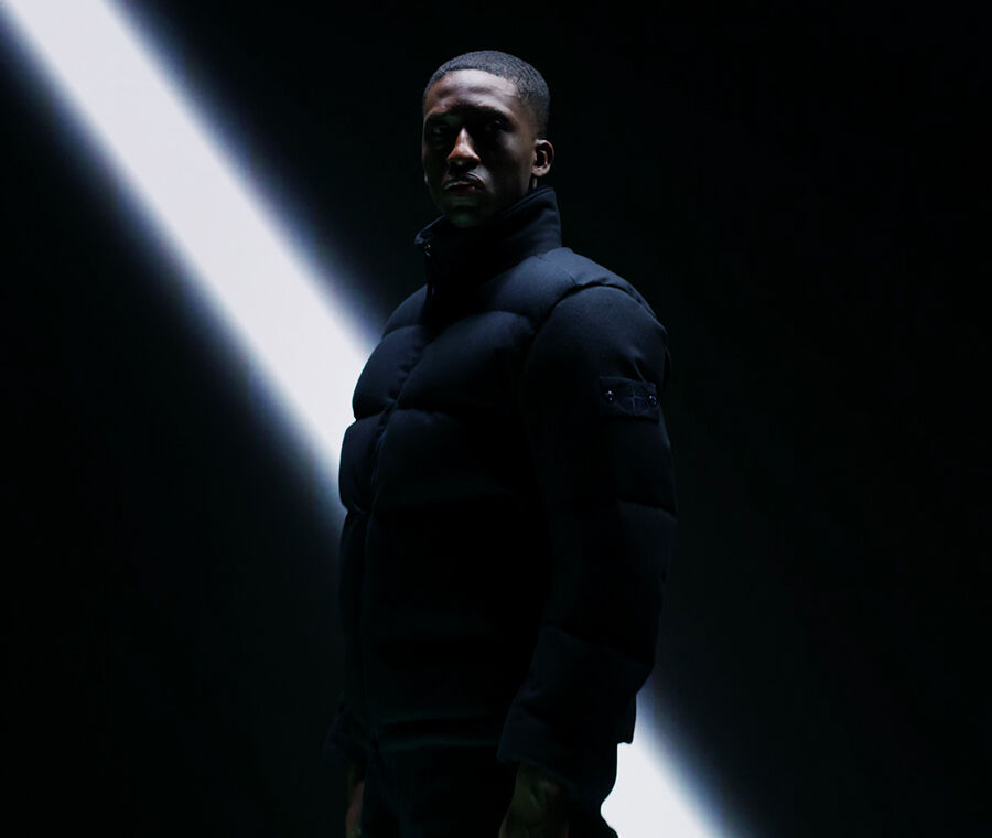 Model wearing a dark jacket in extreme low light, against a black backdrop with one diagonal beam of white light behind him.