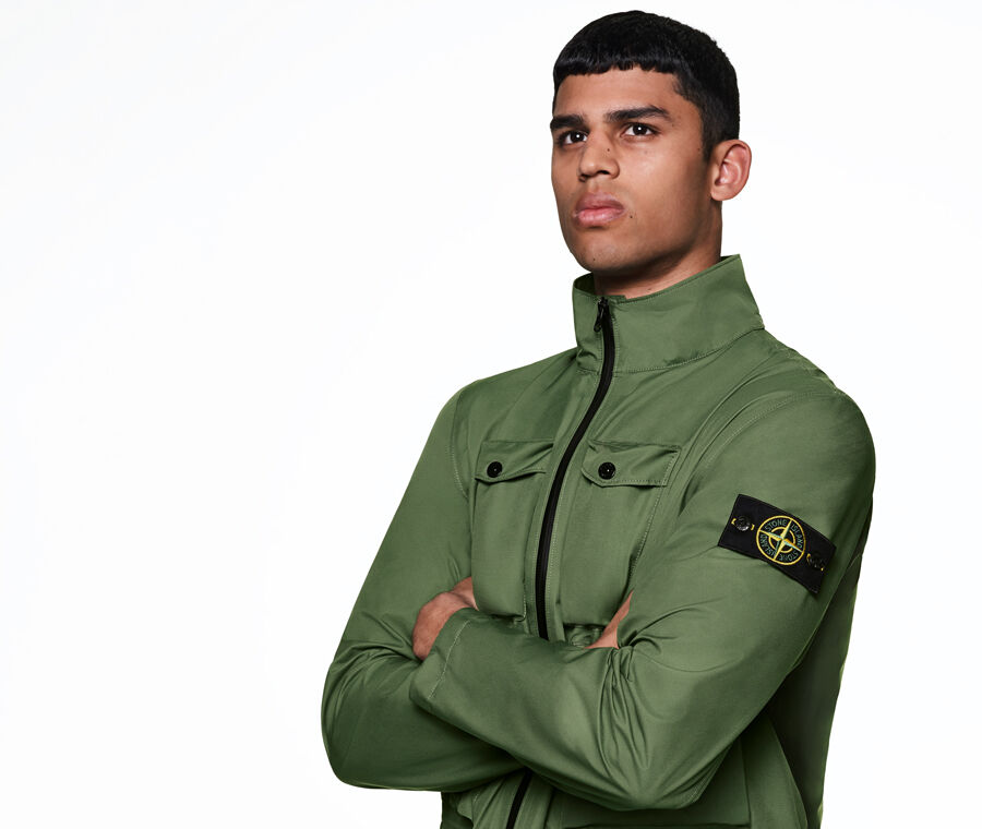 Model posed with crossed arms wearing a zipped green jacket with a high collar, two chest pockets and a Stone Island badge on the sleeve.