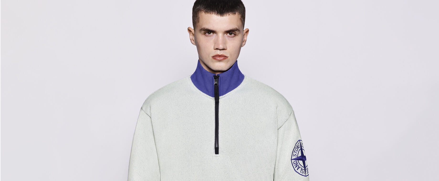 Model wearing an off-white half-zip sweatshirt with a purple neckline and Stone Island logo embroidered on the upper left sleeve.