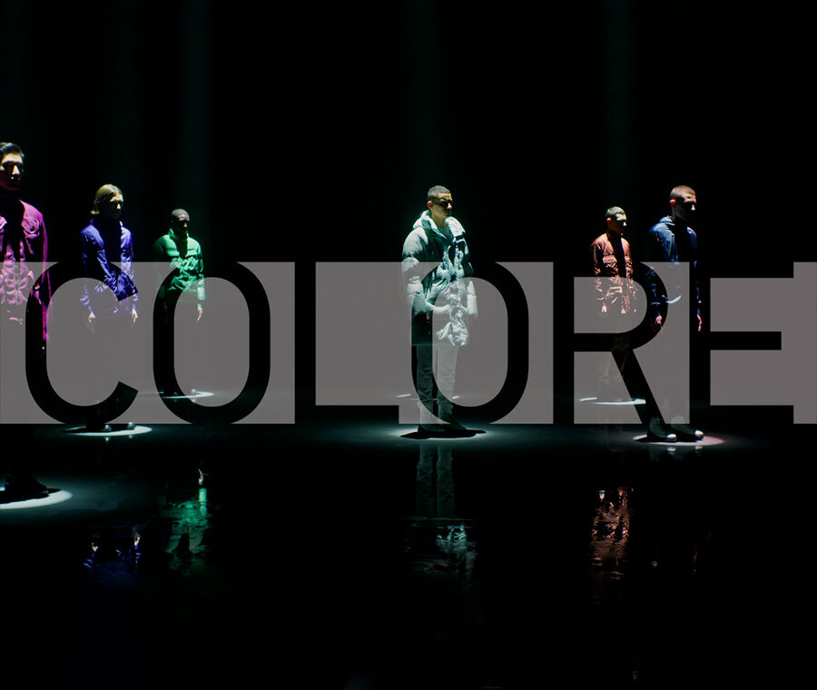 Six male models, each wearing a different colored outfit, stand beneath white spotlights with overlaid text that reads \"Colore\".