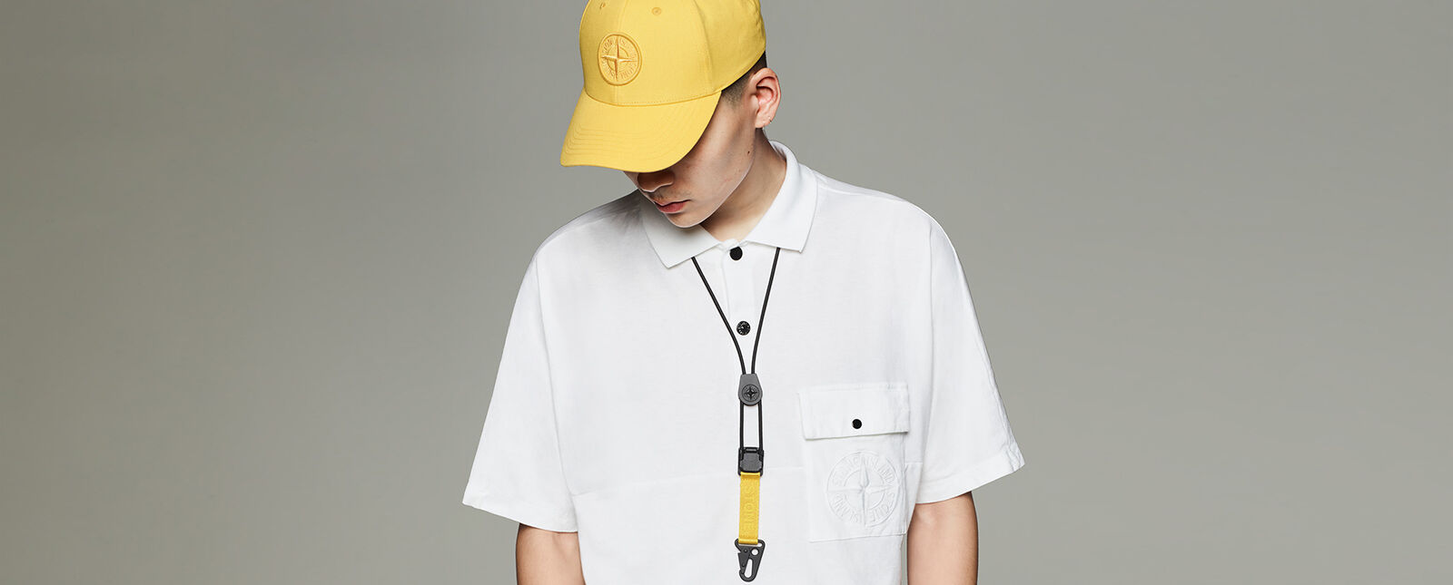 Headshot of a model looking down, wearing a yellow cap with an embroidered compass rose, a white polo and a yellow lanyard around his neck.
