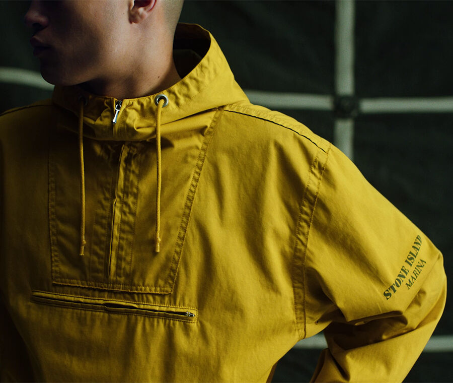 Model in dim lighting wearing yellow hooded quarter-zip jacket with Stone Island Marina logo print on the sleeve.