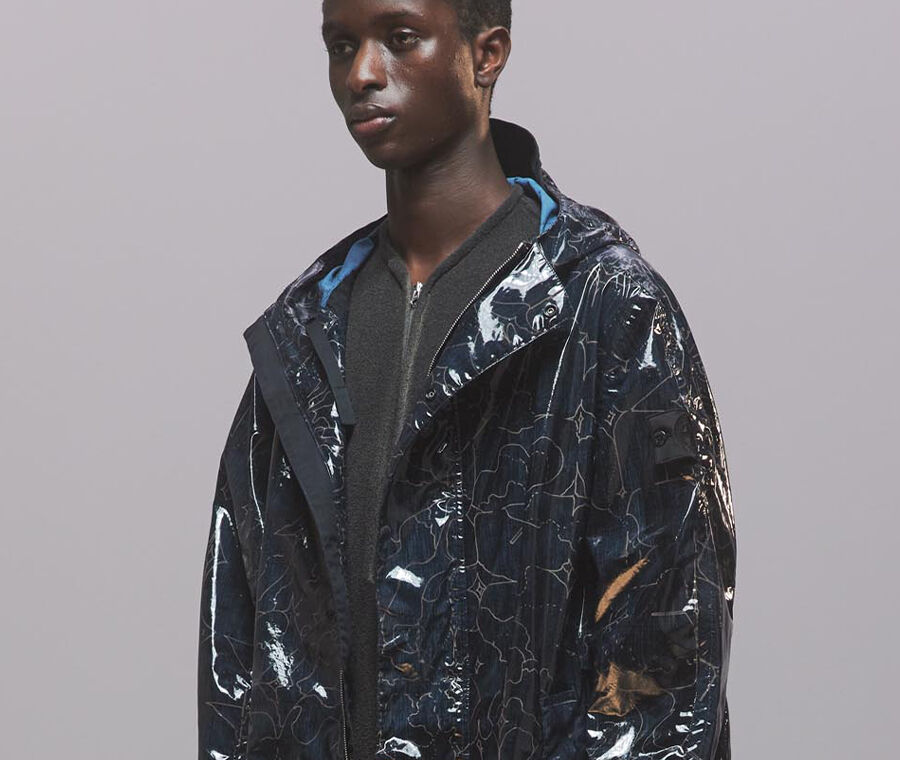 Model wearing navy hooded shiny-effect jacket with all-over abstract motif and Shadow Project badge on sleeve.