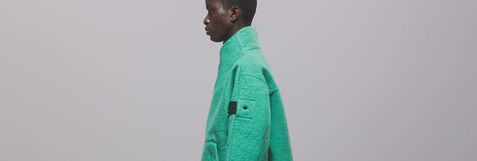 Model, from side, wearing green high-collar fleece jacket with Shadow project badge on sleeve.