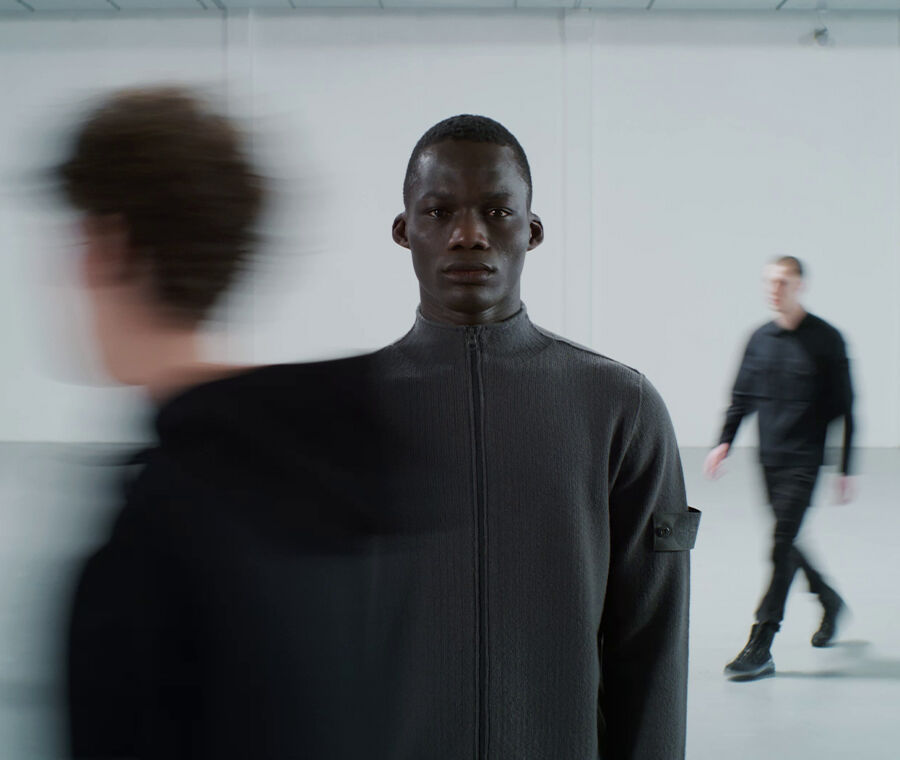 Model wearing gray full-zip sweatshirt with Stone Island Ghost badge on sleeve and timelapse effect.
